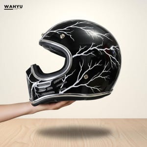 Könnte beinhalten: Schwarzer Motorradhelm mit weißem Blitzmuster. Der Helm hat eine silberne Zierleiste und einen Kinnschutz. Der Helm wird von einer Hand gehalten.