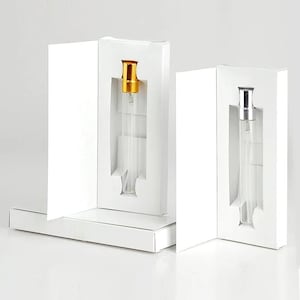 May include: Two clear glass perfume atomizers with gold and silver caps are displayed in white cardboard boxes. The atomizers are slender and cylindrical, designed for fragrance samples or travel. The boxes are open, showcasing the products.