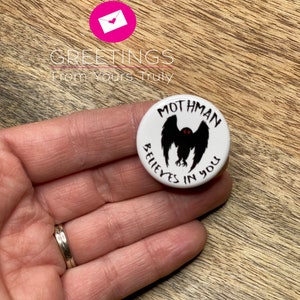 Peut inclure: Un bouton blanc avec un motif de mothman noir et rouge. Le texte sur le bouton est "Mothman Believes in You".