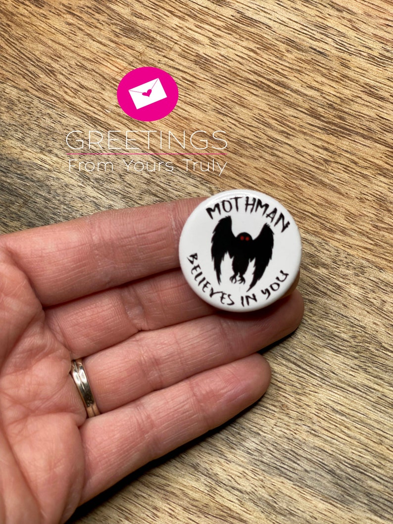 Peut inclure: Un bouton blanc avec un motif de mothman noir et rouge. Le texte sur le bouton est "Mothman Believes in You".