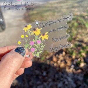 May include: A clear sticker with a quote by Dolly Parton: "Wildflowers don't care where they grow". The sticker features a floral design with yellow wildflowers, green leaves, and two bees.