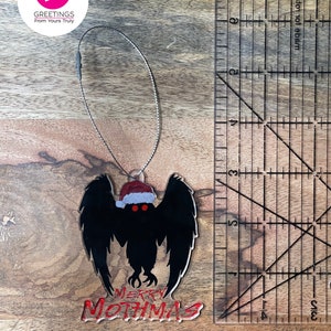May include: A black and red Christmas ornament featuring a silhouette of a mothman wearing a Santa hat with the text "Merry Mothmas".