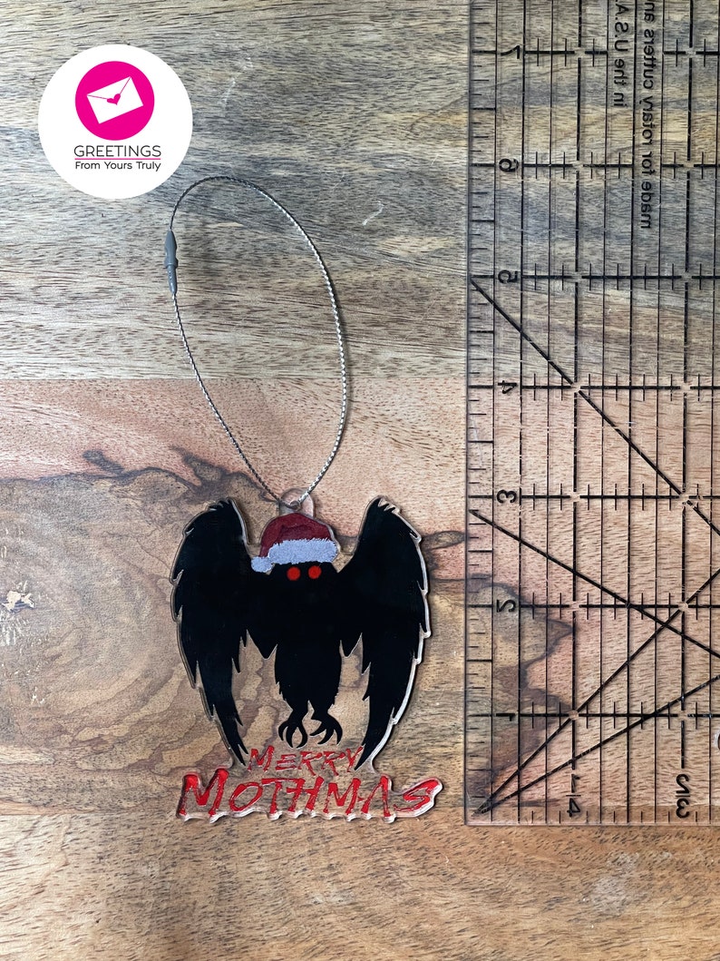 May include: A black and red Christmas ornament featuring a silhouette of a mothman wearing a Santa hat with the text "Merry Mothmas".