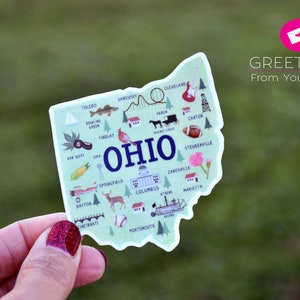 May include: A sticker of the state of Ohio with illustrations of landmarks and cities, including Toledo, Cleveland, Columbus, Cincinnati, and Portsmouth. The sticker is green and white with blue text that says "OHIO". The text "GREETINGS From Yours Truly" is in the upper right corner of the image.