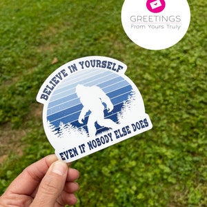 May include: A white sticker with a blue and white sunset background. The sticker features a silhouette of Bigfoot walking through a forest with the text "Believe in Yourself Even if Nobody Else Does".