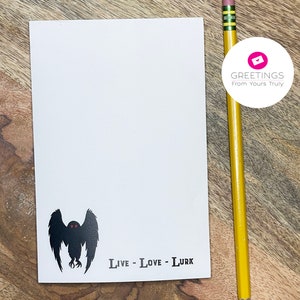 Peut inclure: Une carte blanche avec une silhouette noire d'une cr&eacute;ature aux grandes ailes et aux yeux rouges. Le texte "Live - Love - Lurk" est imprim&eacute; sous la cr&eacute;ature.