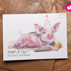 May include: A birthday card featuring a watercolor illustration of a pink pig wearing a party hat and holding a cupcake. The card says "Ham it up! It's your birthday."