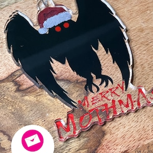 May include: A black and white acrylic ornament of the Mothman with a red Santa hat. The ornament has the text "Merry Mothman" in red.