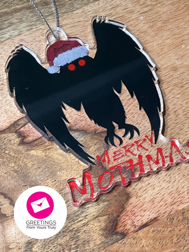 May include: A black and white acrylic ornament of the Mothman with a red Santa hat. The ornament has the text "Merry Mothman" in red.