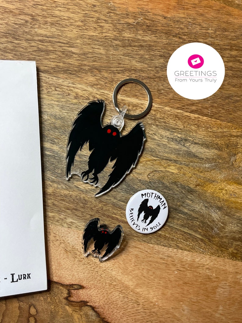 Peut inclure: Un porte-cl&eacute;s Mothman noir et blanc avec des yeux rouges et le texte "Mothman Believes in You". Le porte-cl&eacute;s est attach&eacute; &agrave; un anneau de cl&eacute; argent&eacute;. Il y a aussi une petite &eacute;pingle Mothman noir et blanc avec des yeux rouges.