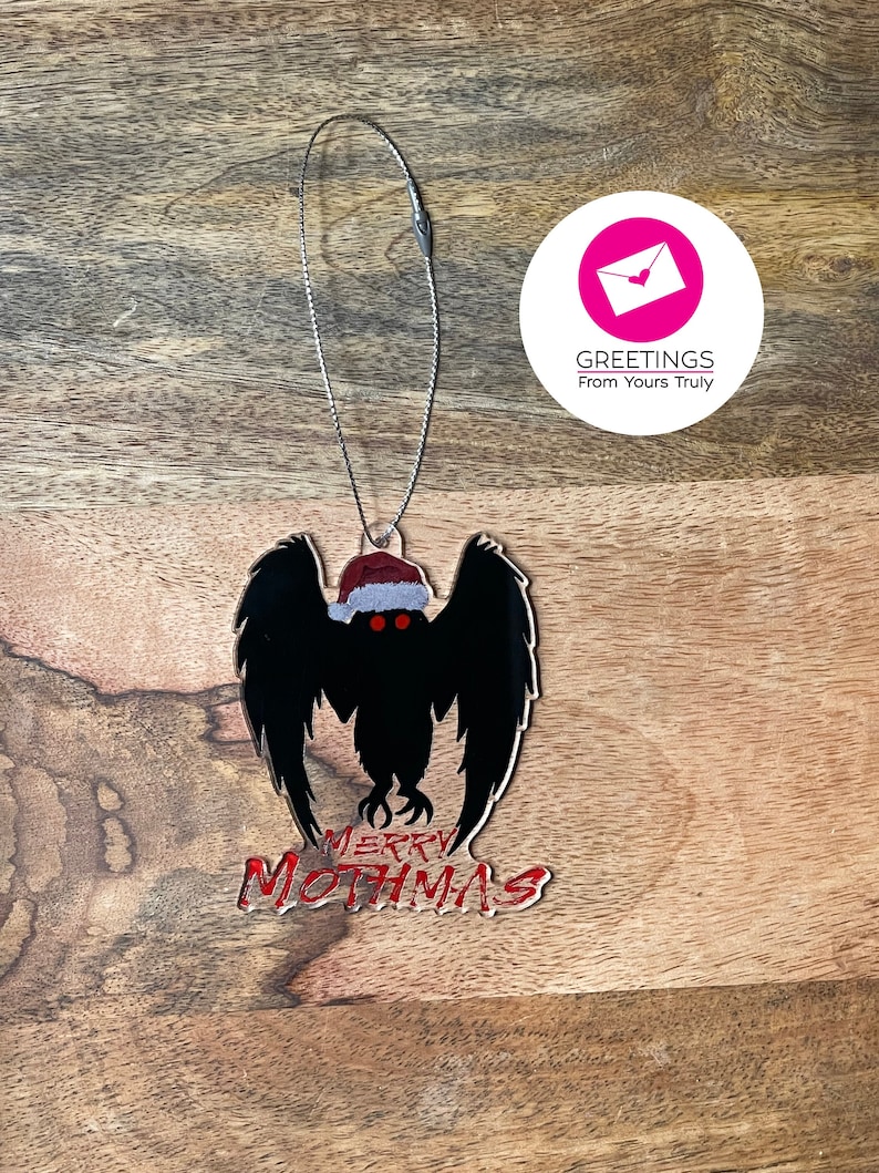 May include: A black mothman ornament with red eyes and a Santa hat. The ornament says "Merry Mothmas" in red lettering.