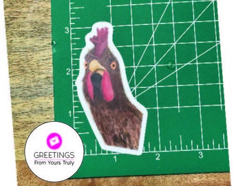 May include: A close-up of a rooster sticker on a green cutting mat. The rooster is brown and white with a red comb and wattle. The sticker is on a white background.