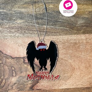 May include: A black mothman Christmas ornament with red eyes and a Santa hat. The ornament says "Merry Mothmas" in red.
