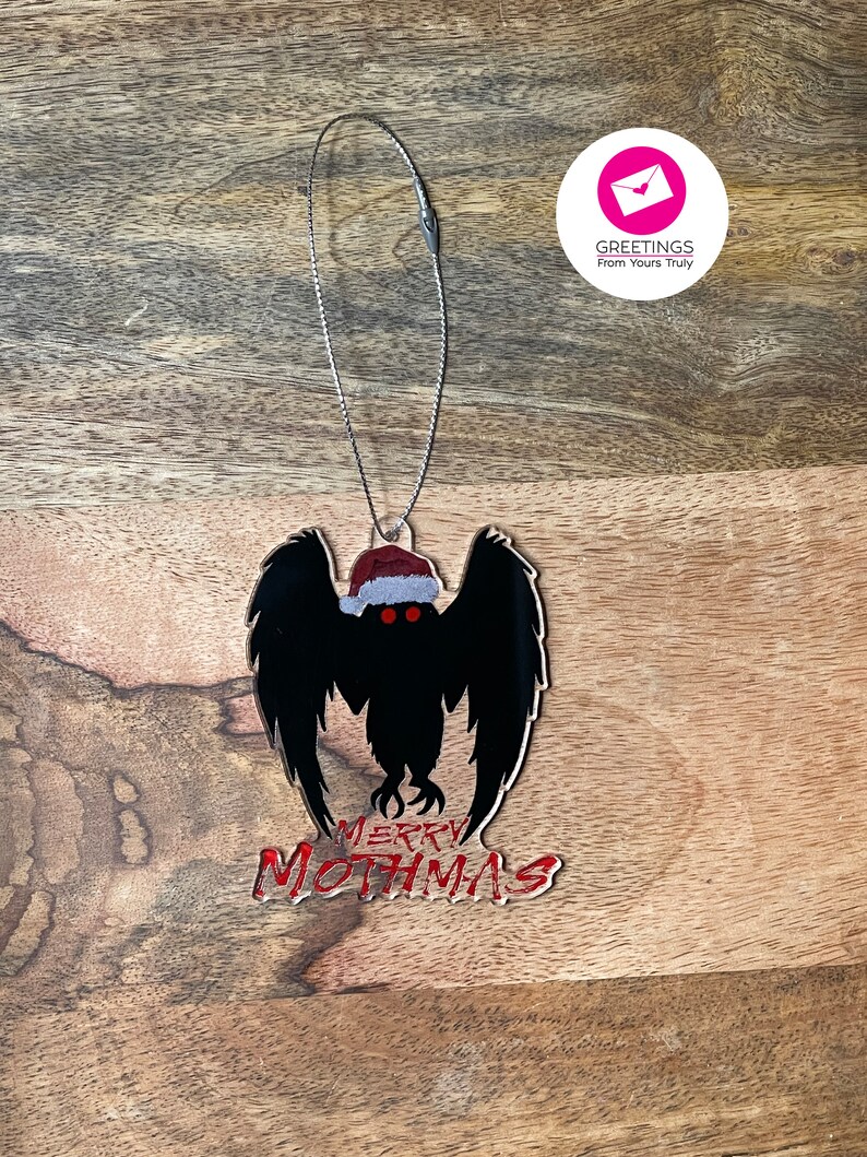 May include: A black mothman Christmas ornament with red eyes and a Santa hat. The ornament says "Merry Mothmas" in red.