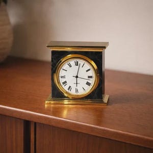May include: A rectangular quartz clock with a black marble face and gold trim. The clock face is white with black Roman numerals and hands. The clock is displayed on a wooden surface.