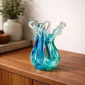 Puede incluir: Un jarrón decorativo de vidrio con una forma orgánica única. El jarrón presenta una mezcla de tonos azules y verdes, creando un efecto acuático arremolinado. Se encuentra sobre una superficie de madera, mostrando su diseño artístico.