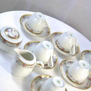 Pode incluir: Um serviço de chá de porcelana branca com detalhes florais e dourados. O conjunto inclui xícaras de chá, pires, um creme e um açucareiro. O padrão floral apresenta flores rosa e verdes. O serviço de chá está disposto sobre uma superfície branca.