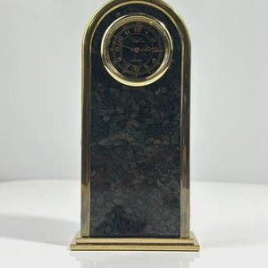 May include: A vintage rectangular clock with a black mosaic face and a gold-coloured frame. The clock face is round with gold hands and numerals. The clock is approximately 18 cm tall and 10 cm wide.