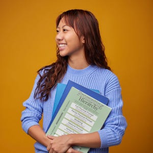 Op de afbeelding: Een persoon in een blauwe trui en jeans houdt een stapel boeken vast. Het bovenste boek is groen en wit met de titel "Prompting Hierarchy" en een lijst met prompts. De achtergrond is oranje.