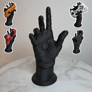 Puede incluir: Un soporte negro con forma de mano para mando de videojuegos. El soporte está diseñado para sujetar un mando de videojuegos. Varios mandos de diferentes colores se muestran en soportes más pequeños. El soporte principal está sobre una superficie blanca.