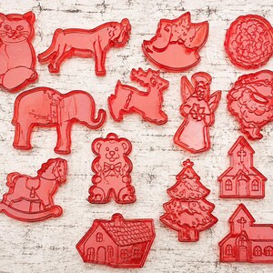 May include: A collection of thirteen red plastic cookie cutters in various shapes, including a cat, a horse, a rocking horse, an elephant, a bear, a reindeer, an angel, a Christmas tree, a Santa Claus, a flower, a house, and two church buildings.