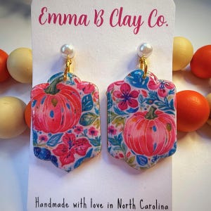 May include: A pair of handmade earrings featuring a floral and pumpkin design. The earrings are rectangular with a scalloped edge, adorned with pink pumpkins and flowers on a white background. The text "Emma B Clay Co." and "Handmade with love in North Carolina" are visible.