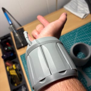 Może przedstawiać: Szary, drukowany w 3D ochraniacz na nadgarstek o cylindrycznym kształcie. Ochraniacz ma pionowe panele i mały, cylindryczny element mocujący. Produkt jest przeznaczony do noszenia na nadgarstku.