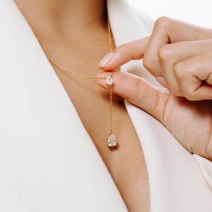May include: A delicate gold necklace with two clear, teardrop-shaped pendants. The necklace is being held by a hand, showcasing the fine details of the jewelry. The background is a white blazer, highlighting the necklace's elegance.