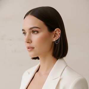 May include: A silver ear cuff with two white pearls and delicate chain detailing. The ear cuff is attached to the ear with a small stud and drapes down the ear with two strands of chain. The jewelry is worn on the ear of a person wearing a white blazer.