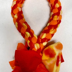 May include: A braided dog toy in shades of orange, yellow, and red. The toy has a loop shape with a large red bow at the base. The plush material is soft and textured, designed for playful interaction.