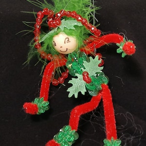 May include: A whimsical Christmas ornament shaped like a small elf. The elf has a wooden face with painted features, green feather hair, and a red pipe cleaner body. Green and red beaded accents adorn the elf's outfit, creating a festive holiday decoration.