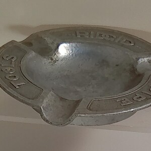 Puede incluir: Un cenicero ovalado vintage de metal plateado. El cenicero tiene las palabras "PIPE" y "TOOLS" en relieve en los lados, con "RIDGID" en el centro. Cuenta con una peque&ntilde;a muesca para apoyar los cigarrillos.