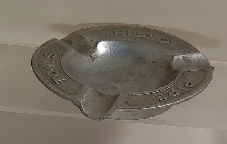 Puede incluir: Un cenicero ovalado vintage de metal plateado. El cenicero tiene las palabras "PIPE" y "TOOLS" en relieve en los lados, con "RIDGID" en el centro. Cuenta con una peque&ntilde;a muesca para apoyar los cigarrillos.