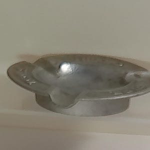 Puede incluir: Un cenicero plateado con un dise&ntilde;o circular y una peque&ntilde;a base elevada. El cenicero tiene una forma curva con un borde y una muesca para apoyar los cigarrillos. El cenicero es de metal.
