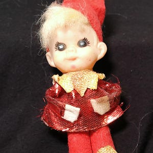 May include: A vintage Christmas elf doll with a red felt hat and outfit, accented with gold trim. The doll has a painted face with blue eyes and blonde hair. A classic holiday decoration.