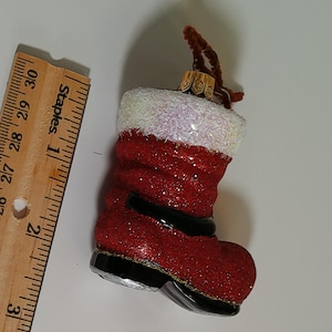 May include: A festive Christmas ornament shaped like a red Santa boot with glitter. The boot has a white, sparkly cuff and a black strap. A gold-colored metal cap and brown pipe cleaner are at the top.