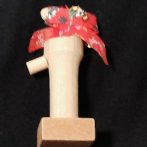May include: A wooden bottle stopper with a red fabric top. The stopper has a T-shape with a rectangular base and a cylindrical body. The red fabric has a white pattern.