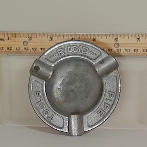 Puede incluir: Un cenicero circular plateado con la marca "RIDGID PIPE TOOLS" grabada alrededor del borde. El cenicero tiene tres apoyos para cigarrillos y un cuenco central. Una regla est&aacute; colocada junto al cenicero.