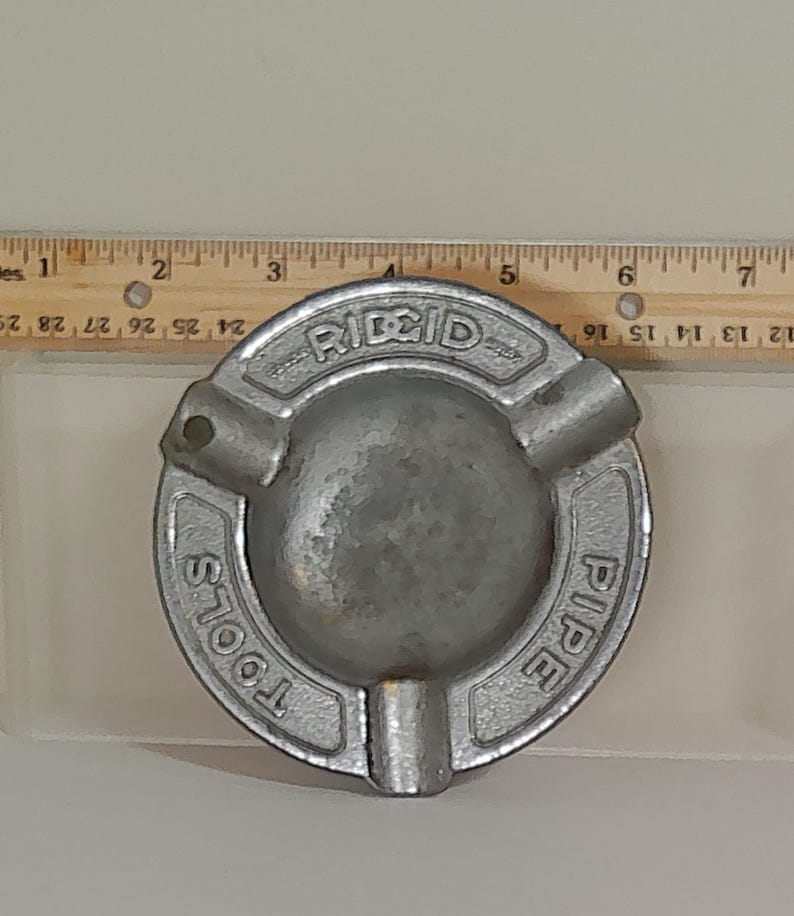 Puede incluir: Un cenicero circular plateado con la marca "RIDGID PIPE TOOLS" grabada alrededor del borde. El cenicero tiene tres apoyos para cigarrillos y un cuenco central. Una regla est&aacute; colocada junto al cenicero.