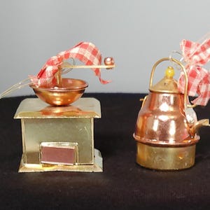May include: Two miniature kitchen ornaments. One is a copper-colored stove with a bowl and a red and white checkered ribbon. The other is a copper-colored teapot with a yellow knob and a red and white checkered ribbon.