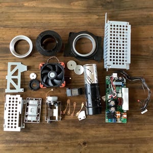 May include: A collection of disassembled electronic components including a black cooling fan, a green circuit board with attached wires, a perforated metal cage, various plastic rings, a cylindrical housing, and small metal brackets arranged on a surface.