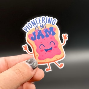 May include: A colorful sticker featuring a smiling piece of toast with jam. The toast has arms and legs and the text "Pioneering is my Jam" in a blue font. The sticker is on a black background.