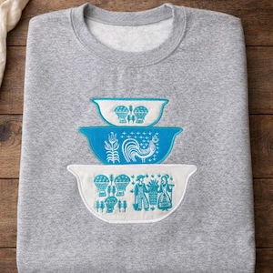 以下が含まれることがあります： 青と白の刺繍が施された3つのボウルが特徴のグレーのスウェットシャツ。ボウルには、雄鶏、熱気球、人物など、さまざまなデザインがあります。スウェットシャツは木製の表面に折りたたまれています。