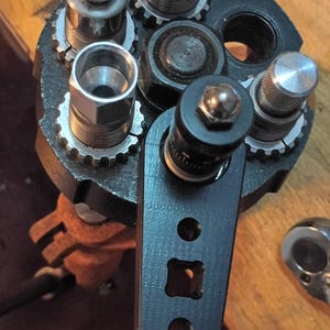 May include: A black metal turret press head featuring multiple circular die stations with silver steel components. A black 3D-printed adjustment arm with a central dial and square cutout is attached to one station, surrounded by various threaded metal parts.