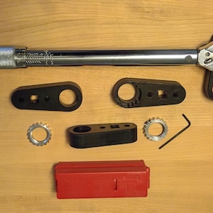 May include: A silver metal torque wrench with a knurled handle, three black plastic adapters, two silver metal serrated rings, a small L-shaped hex key, and a rectangular red plastic storage case arranged on a flat surface.