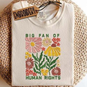 Peut inclure: T-shirt crème avec un motif floral brodé et le texte "BIG FAN OF HUMAN RIGHTS" en vert. Le motif présente des fleurs roses, jaunes et rouges avec des feuilles vertes. Une étiquette avec le mot "EMBROIDERY" est attachée au t-shirt.