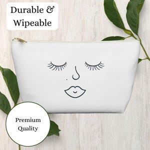Könnte beinhalten: Weiße Kosmetiktasche mit goldenem Reißverschluss, mit einem minimalistischen Gesichtsdesign mit geschlossenen Augen, einer Nase und schwarzen Lippen. Die Tasche trägt die Aufschrift "Durable & Wipeable" und "Premium Quality".