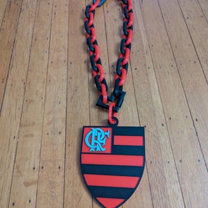May include: A necklace featuring a large pendant with a shield design in red and black stripes, with a teal emblem in the upper left corner. The necklace has a chain made of alternating red and black links.