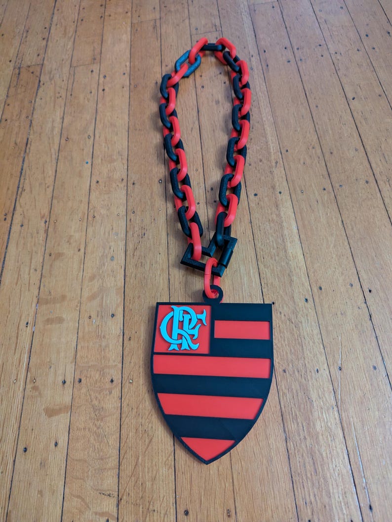 May include: A necklace featuring a large pendant with a shield design in red and black stripes, with a teal emblem in the upper left corner. The necklace has a chain made of alternating red and black links.