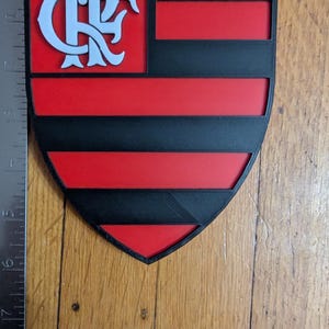 May include: A shield-shaped emblem featuring the Flamengo logo in white on a black and red background. The design includes the team's initials and horizontal stripes, with a small loop at the top for hanging.